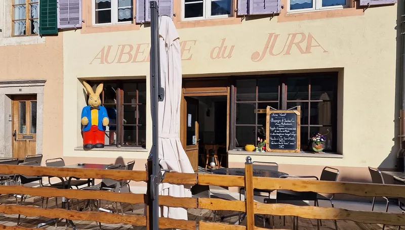 Die Fassade der Auberge du Jura: der auf den Verputz gemalte Name, lavendelfarbene Fensterläden, die Holzterrasse und die Tafel mit dem Tagesmenü.