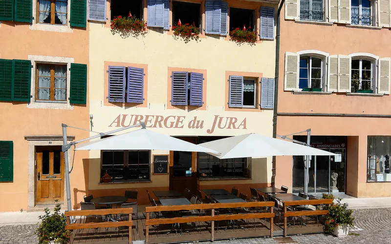 La façade de l'Auberge du Jura : le nom peint sur le crépi, les volets lavande, les géraniums aux fenêtres et la terrasse abritée sous ses parasols.