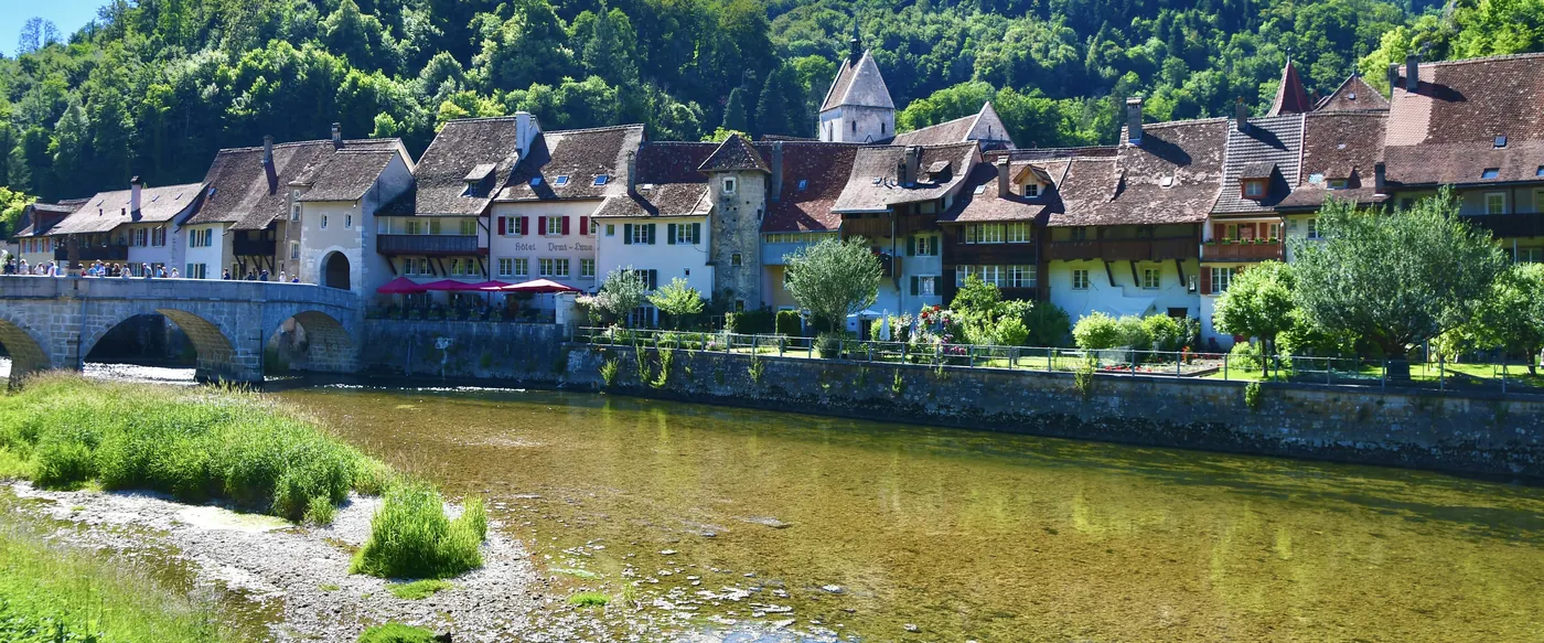 La vieille ville de Saint-Ursanne vue depuis le Doubs : le pont de pierre, les toits de tuiles, le clocher de la collégiale et les collines boisées.