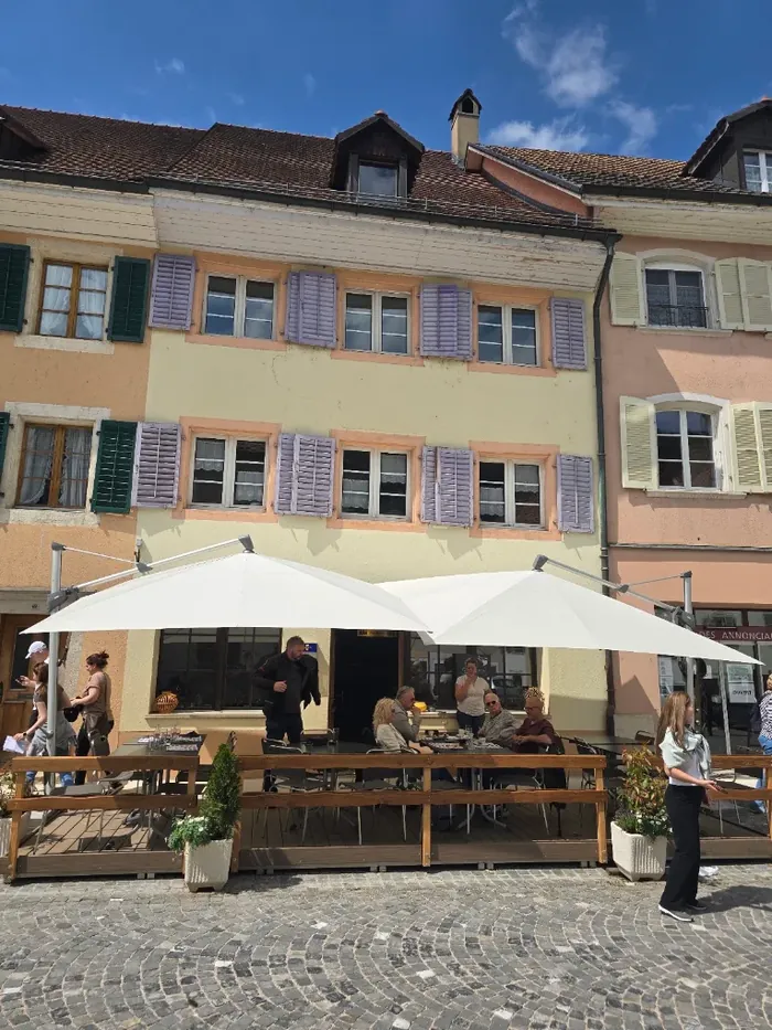 La façade jaune pâle aux volets lavande de l'Auberge du Jura et sa terrasse abritée par deux grands parasols blancs.